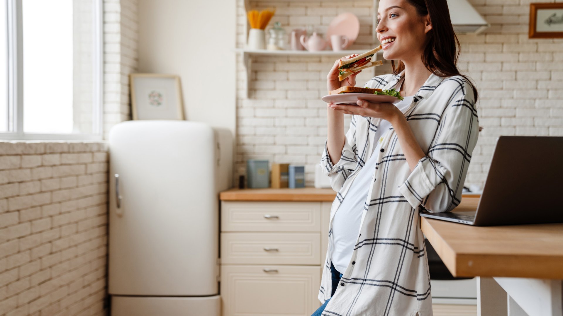 Kobieta w ciazy je kanapke opierajac sie o blat stolu