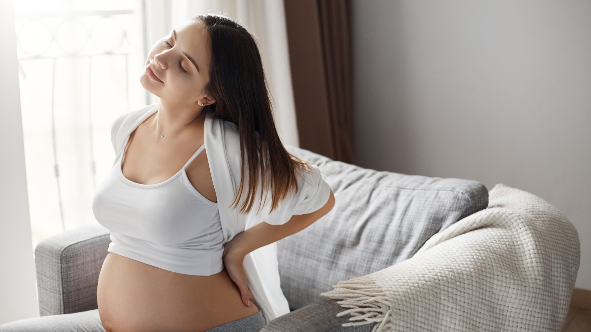 Kobieta w ciazy trzyma sie za bolace plecy siedzac na fotelu