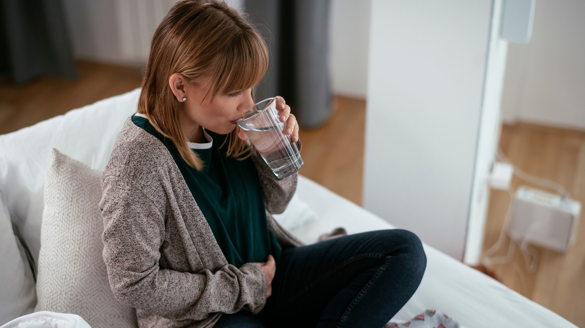 Kobieta w ciazy pije wode na sofie trzymajac się za brzuch