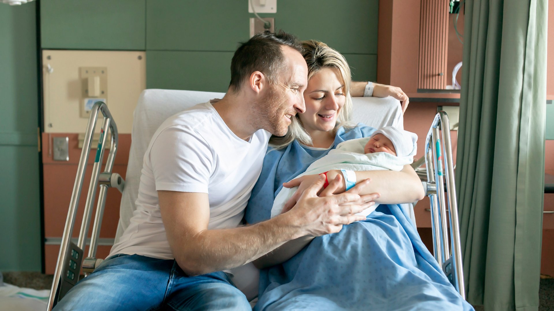 Rodzice przygladaja sie noworodkowi siedzac na szpitalnym lozku
