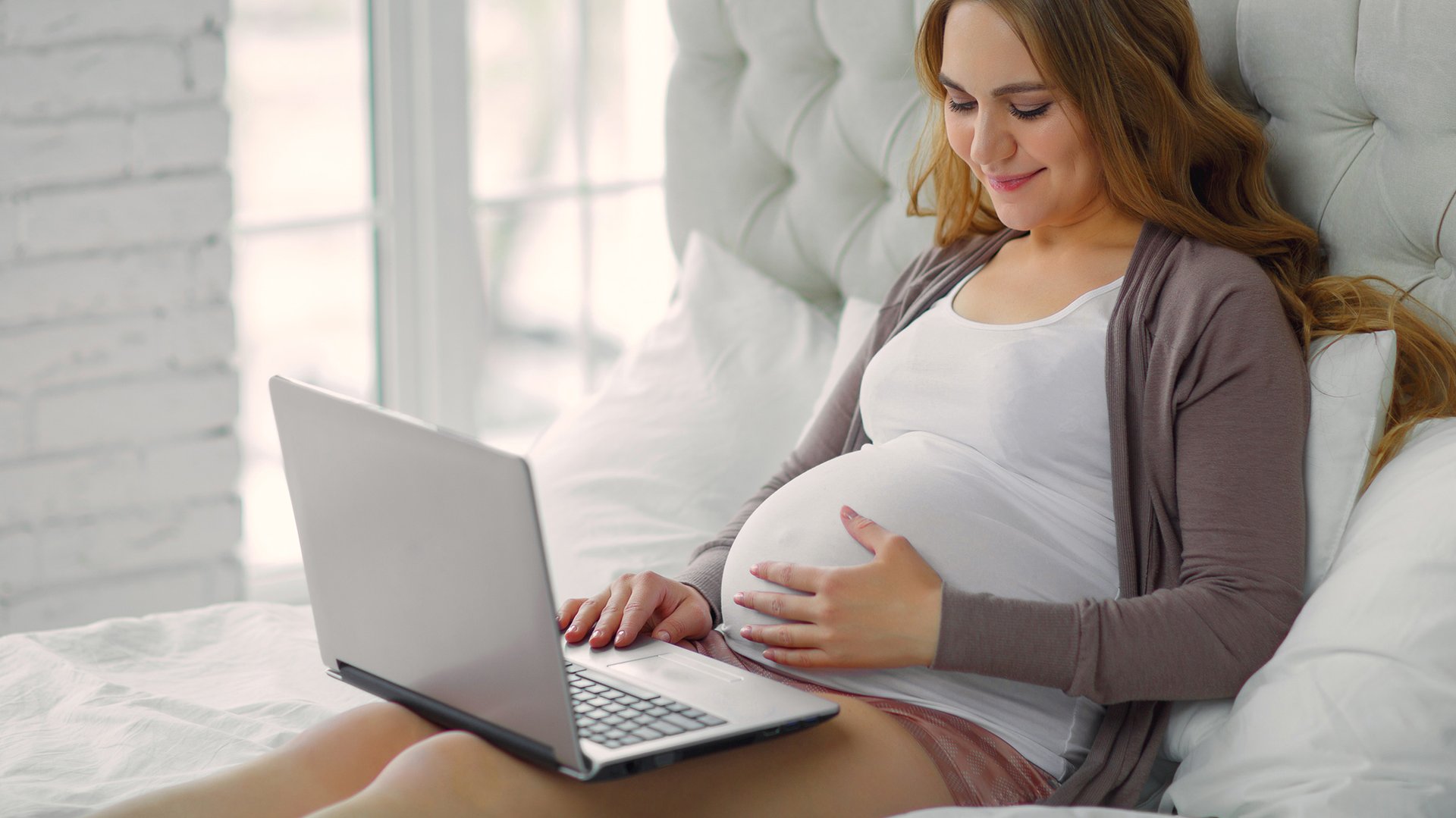 Kobieta w ciazy trzyma laptopa na nogach lezac na lozku